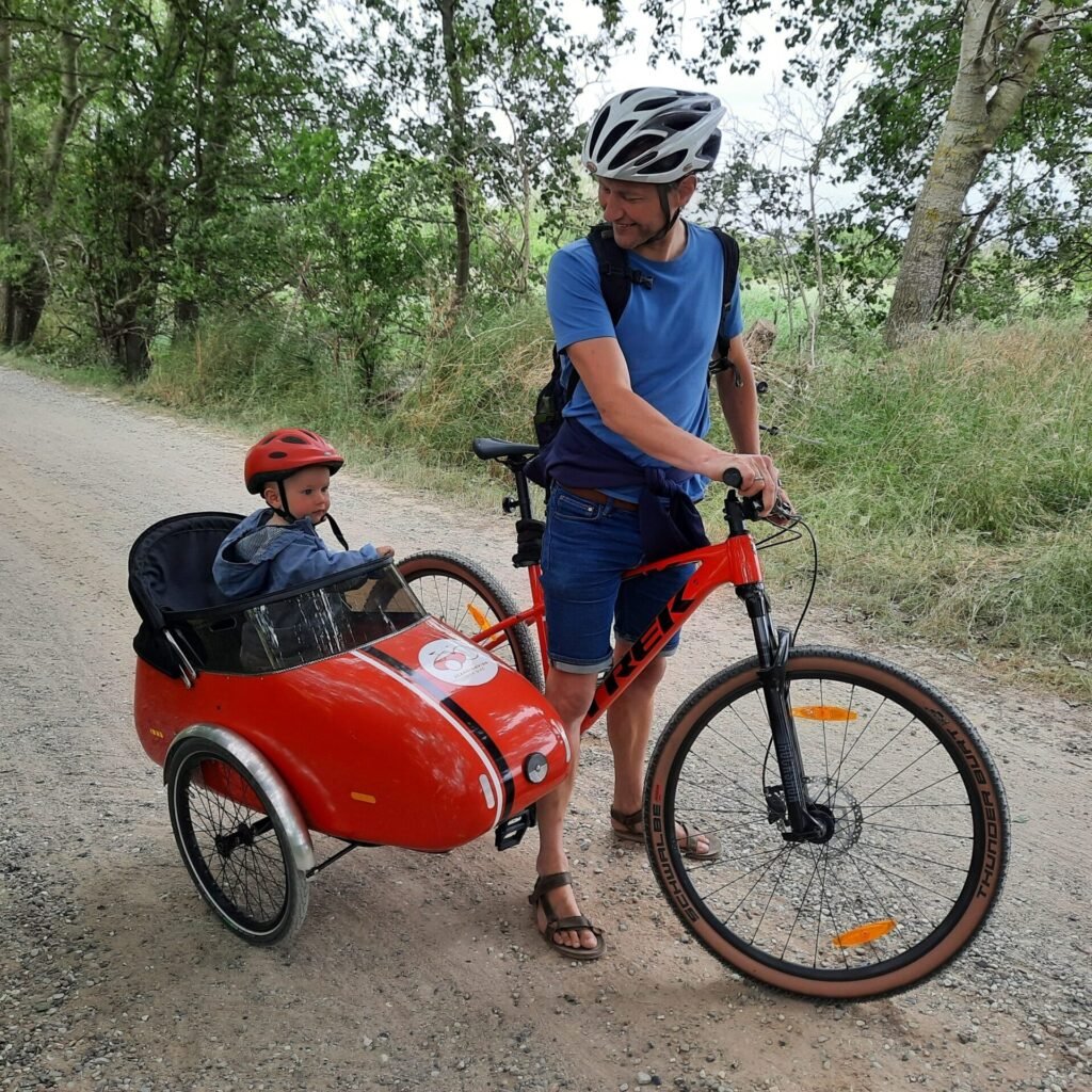 Een kindje in een rode Scandinavian Sidebike aan een mountainbike op een gravelpad.